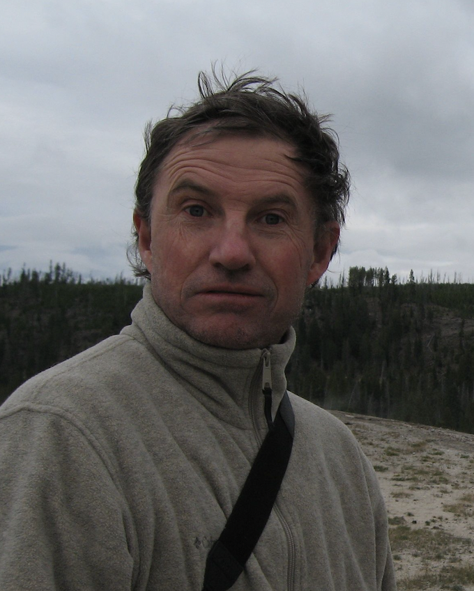 A man in a beige fleece jacket outdoors with a cloudy sky and forested hillside in the background.