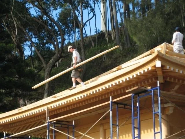 Two men working on the roof of a wooden building, one walking with a long wooden plank, the other standing on the roof, surrounded by scaffolding and trees.