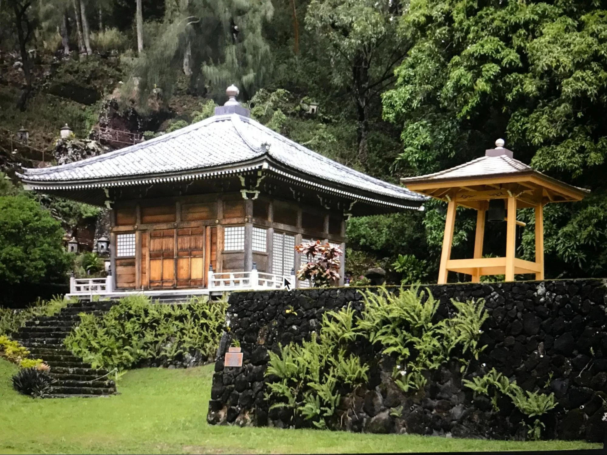 Traditional Japanese temple with wooden structure, tiled roof, surrounded by greenery, elevated on a stone foundation, with stone steps leading up to it, and a small wooden pagoda-style structure nearby.