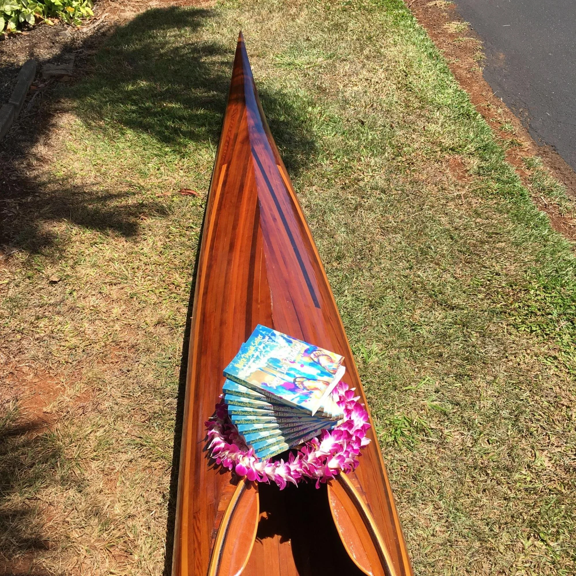 Pointer of a wooden canoe with a stack of magazines or books on a bed of pink and white flowers, on grass beside a paved road.