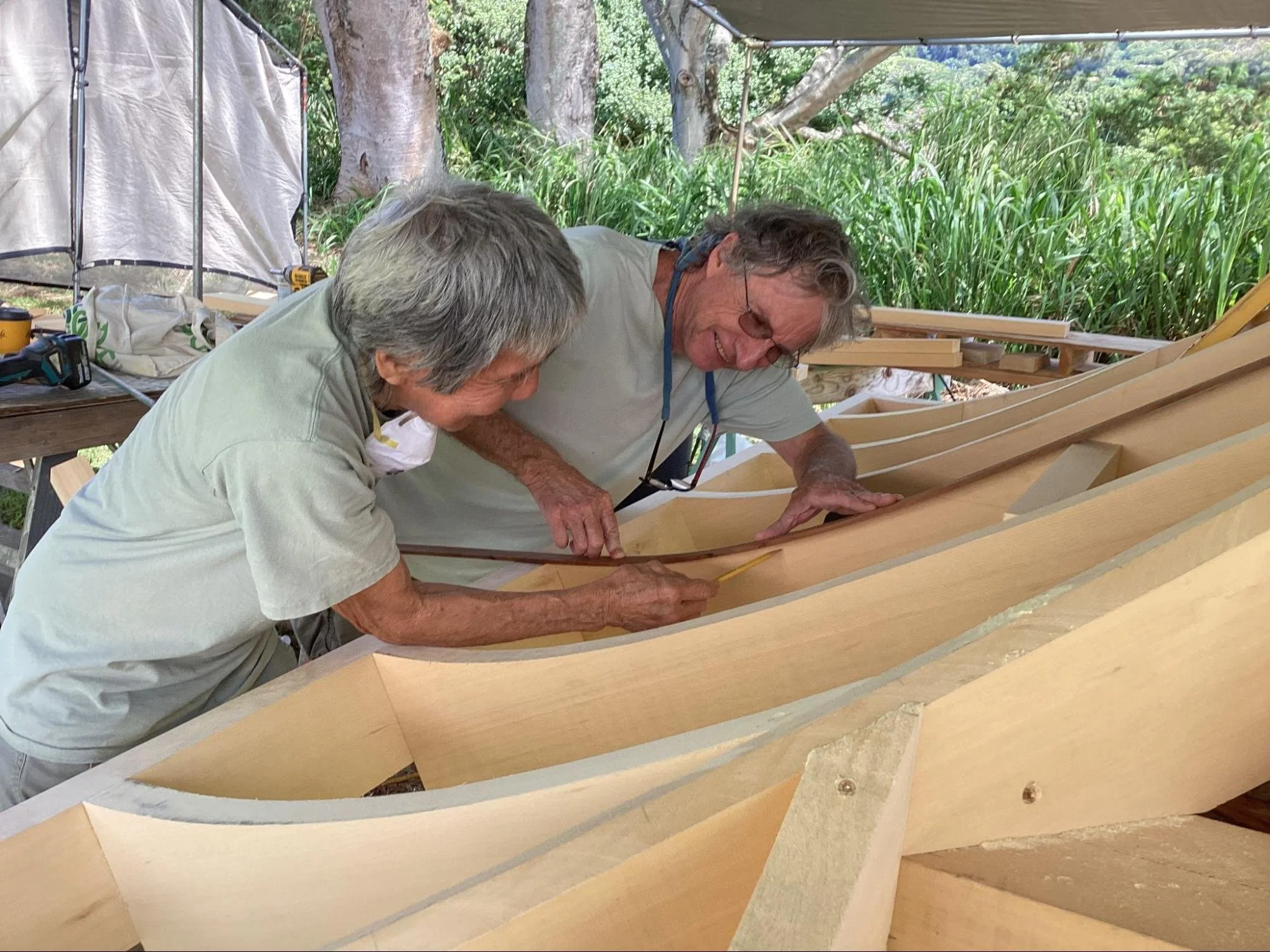 Two older men working on a wooden boat or structure outdoors, surrounded by green trees and plants, under a canopy or tent.