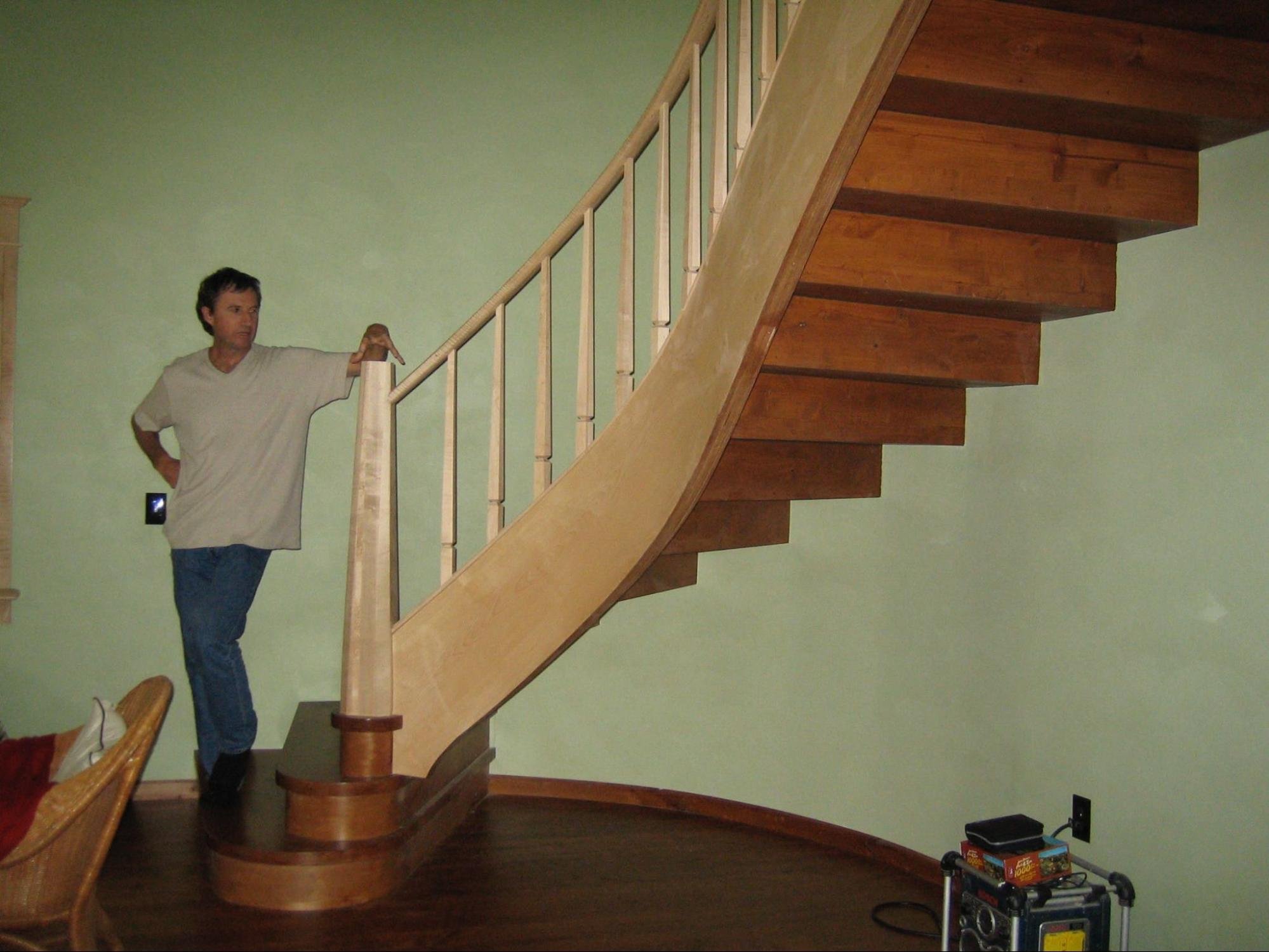 A man in a white T-shirt and jeans is standing at the bottom of a wooden staircase, pointing upward. The staircase has wooden steps and a light-colored handrail, leading to the second floor, with a green wall in the background and dark hardwood flooring on the first floor. There are also some objects, including a black device and a small cart, near the wall.