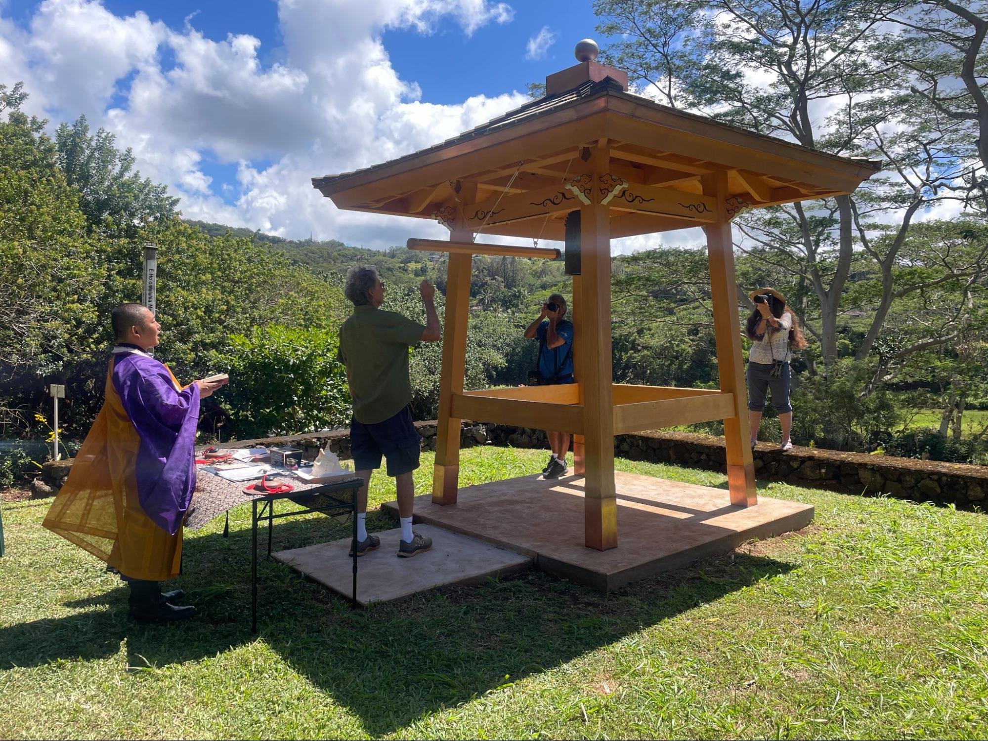 People gathered around a small, traditional Japanese-style wooden shrine outdoors on a bright, partly cloudy day. Some individuals appear to be taking photographs or inspecting the structure, with lush green trees and distant hills in the background.