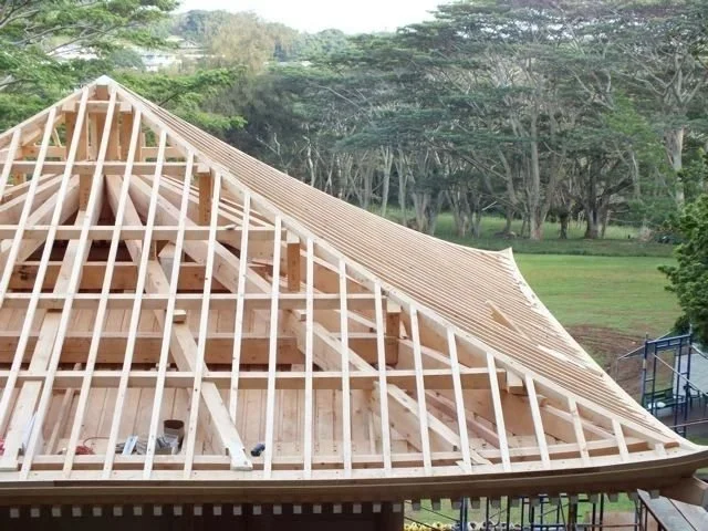 Wooden framework of a roof under construction in a park with trees in the background.