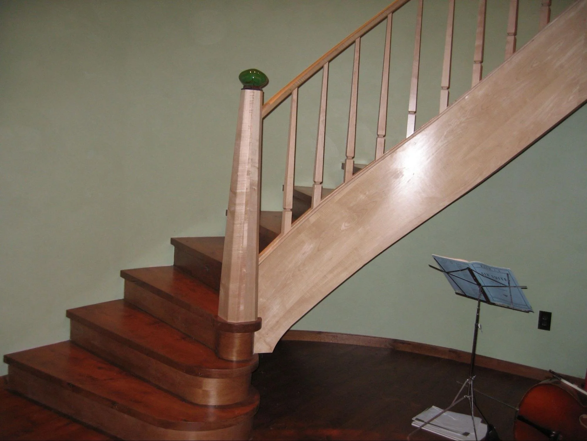 Wooden staircase with a green glass decorative piece on the newel post, against a green wall, with a music stand nearby.