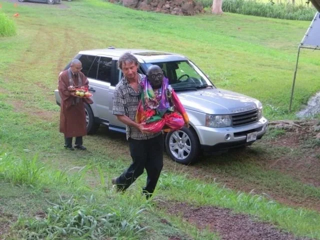 Two people walking in a grassy area near a silver SUV, with an older person carrying a red bag and wearing colorful clothing, and another person in the background holding a tray of fruit, all outdoors.