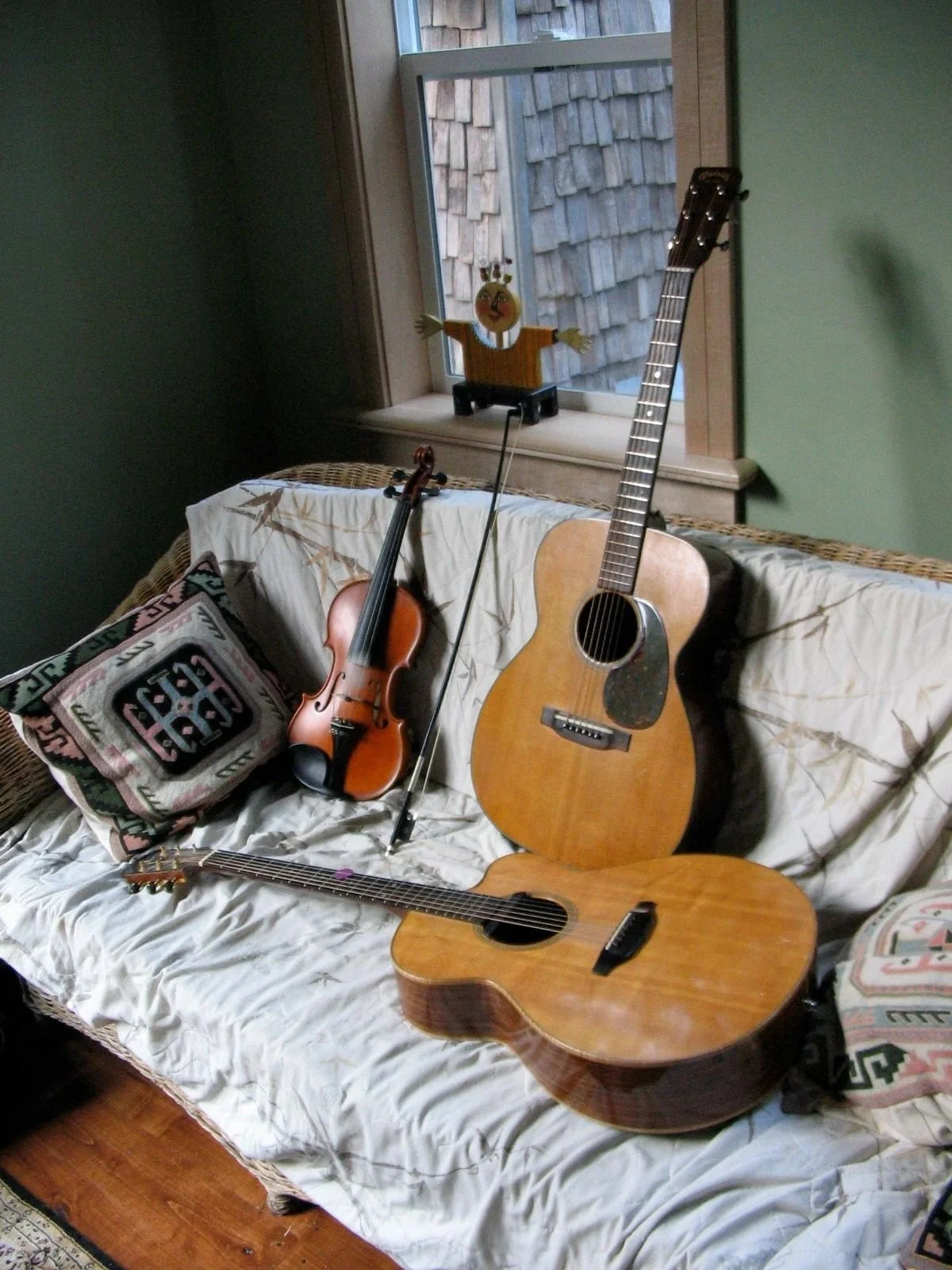 A cozy indoor scene with two guitars, a violin, and a pillow on a sofa. A window with a doll on the windowsill in the background.