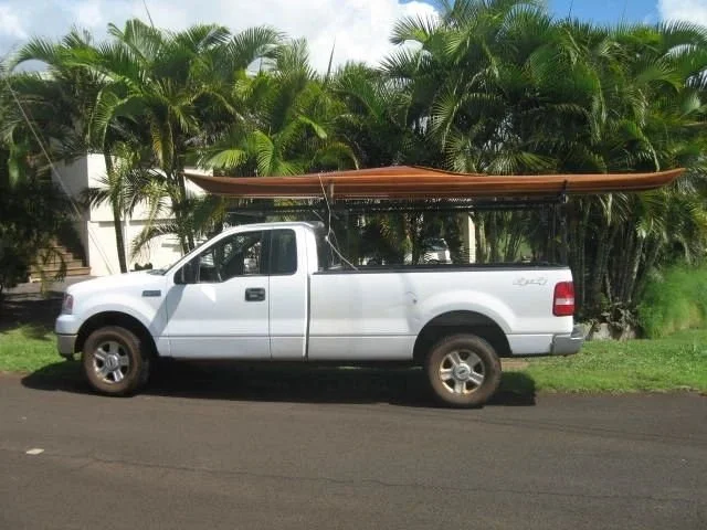 A white pickup truck parked on a street with a brown boat on top, surrounded by tropical palm trees.