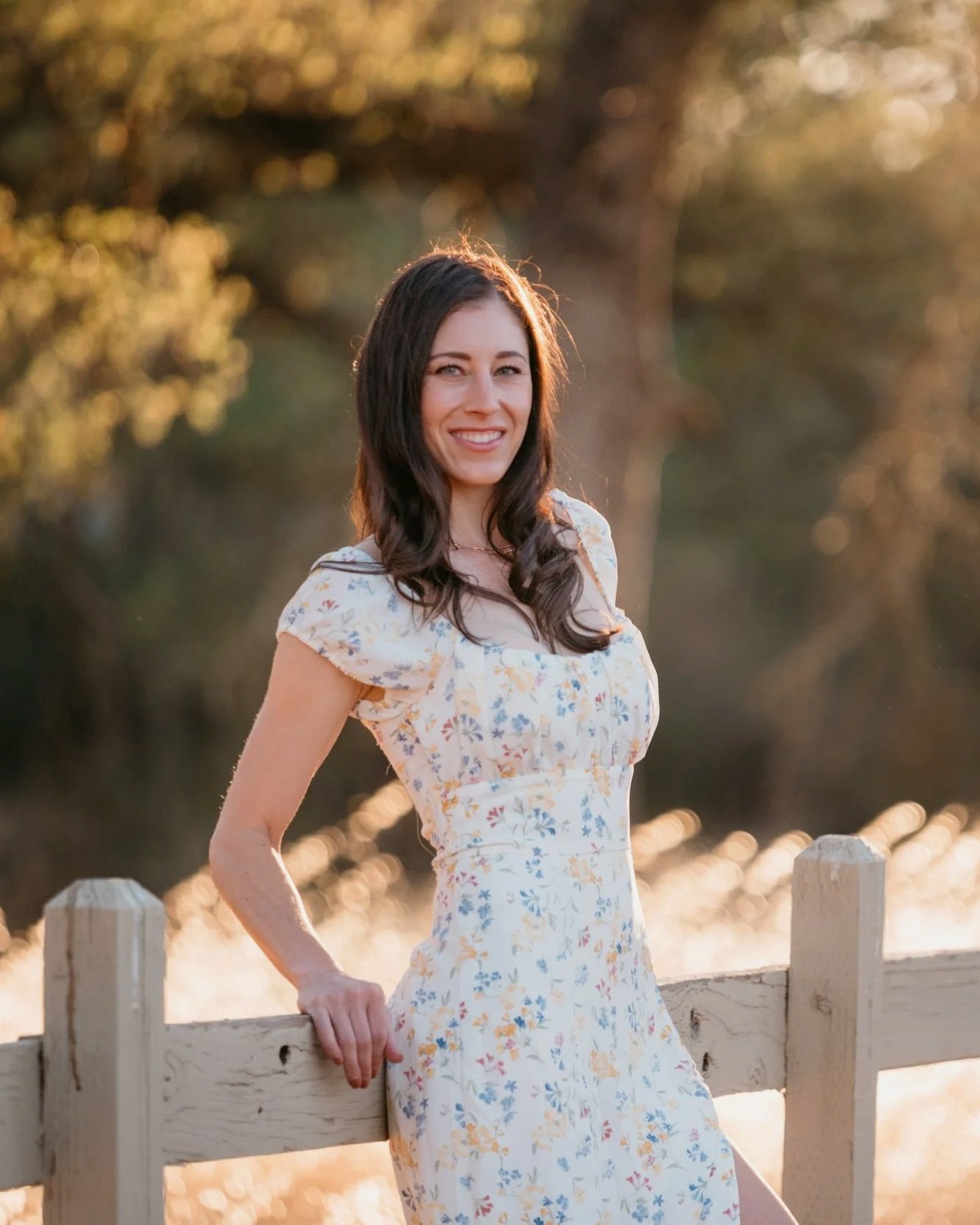 A woman with dark brown hair, smiling, in a yellow floral dress, standing outdoors near a wooden fence with trees and water in the background, during sunset.