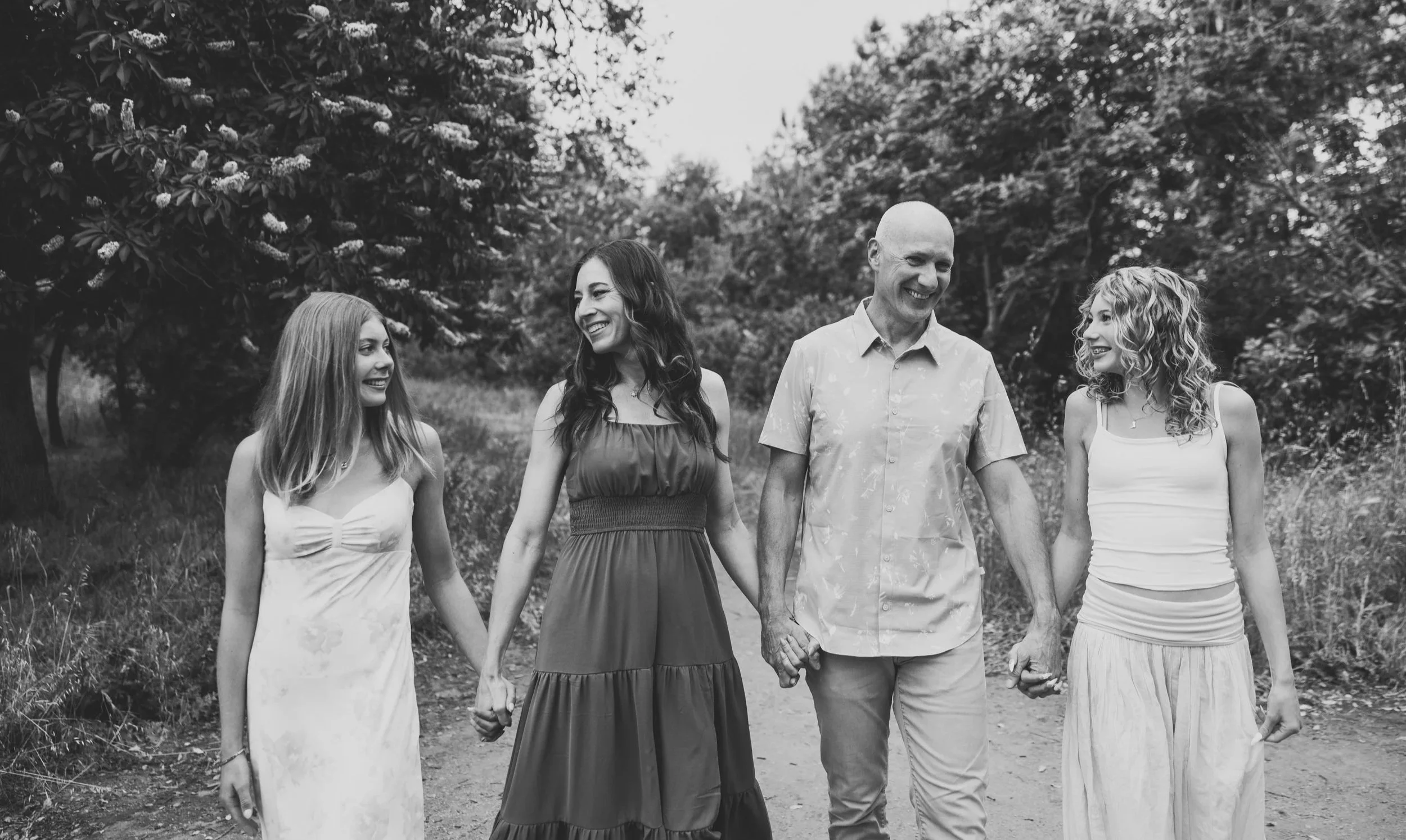 Three women and one man walking in a park, holding hands, smiling, surrounded by trees, black and white photo.