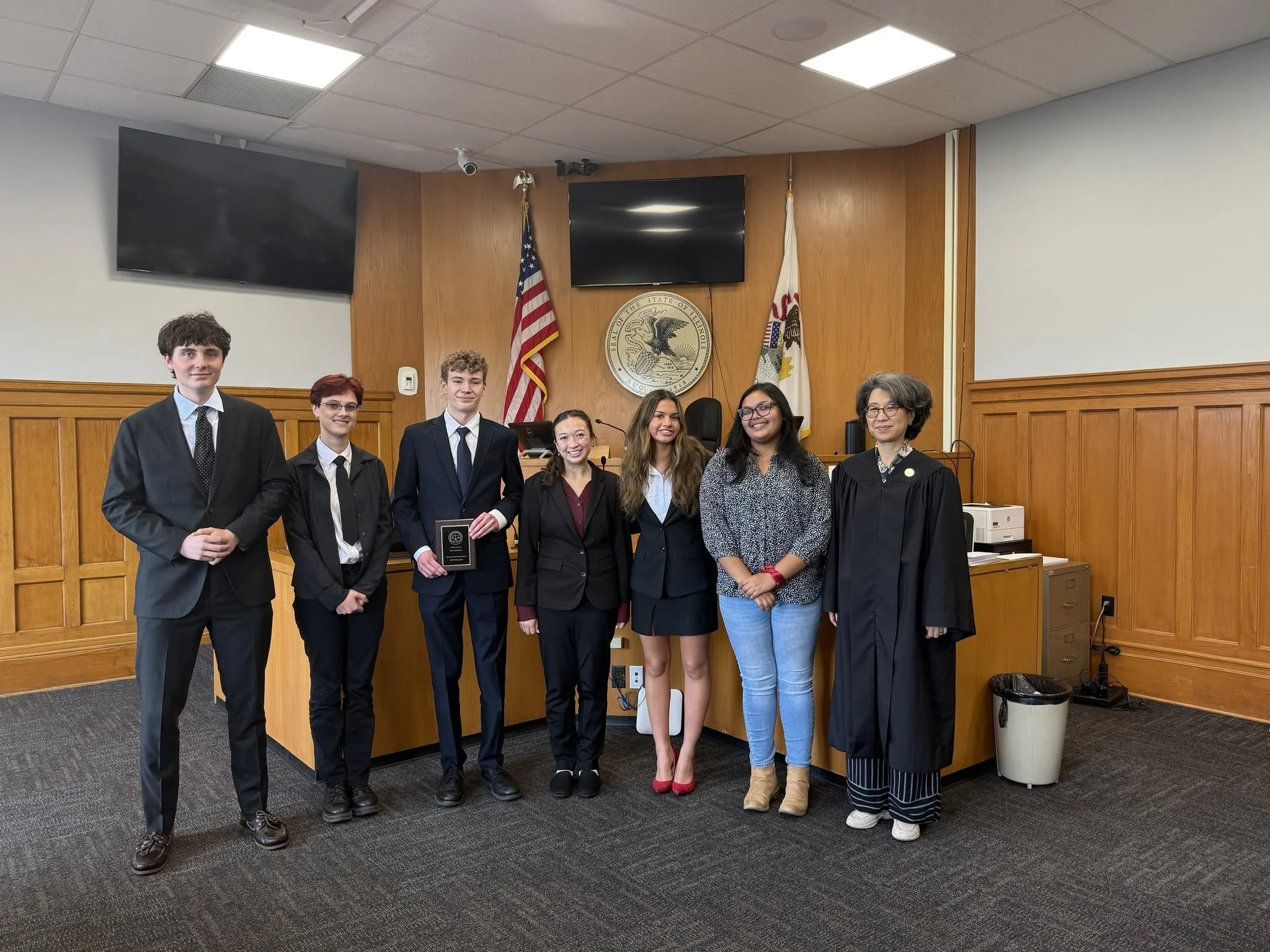 A group of seven diverse young adults and a woman judge standing in a courtroom with wood-paneled walls, flags, and a seal of Illinois in the background.