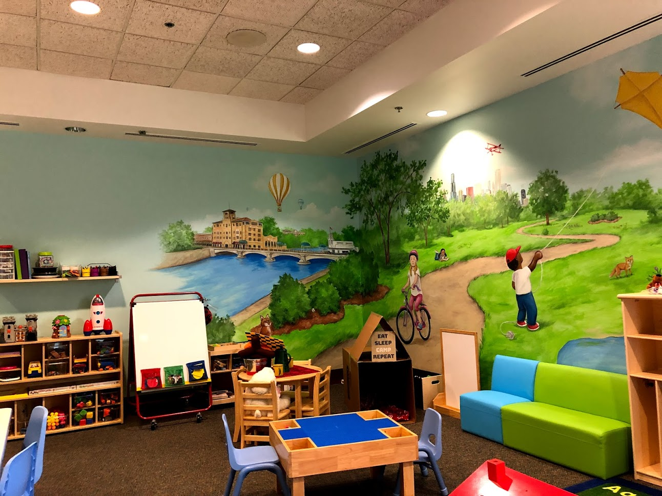 Colorful children’s playroom with a mural of a park scene, featuring a bridge over a river, trees, a girl riding a bike, a boy flying a kite, and animals. There are chairs, shelves with toys, a green and blue sofa, and a small table.