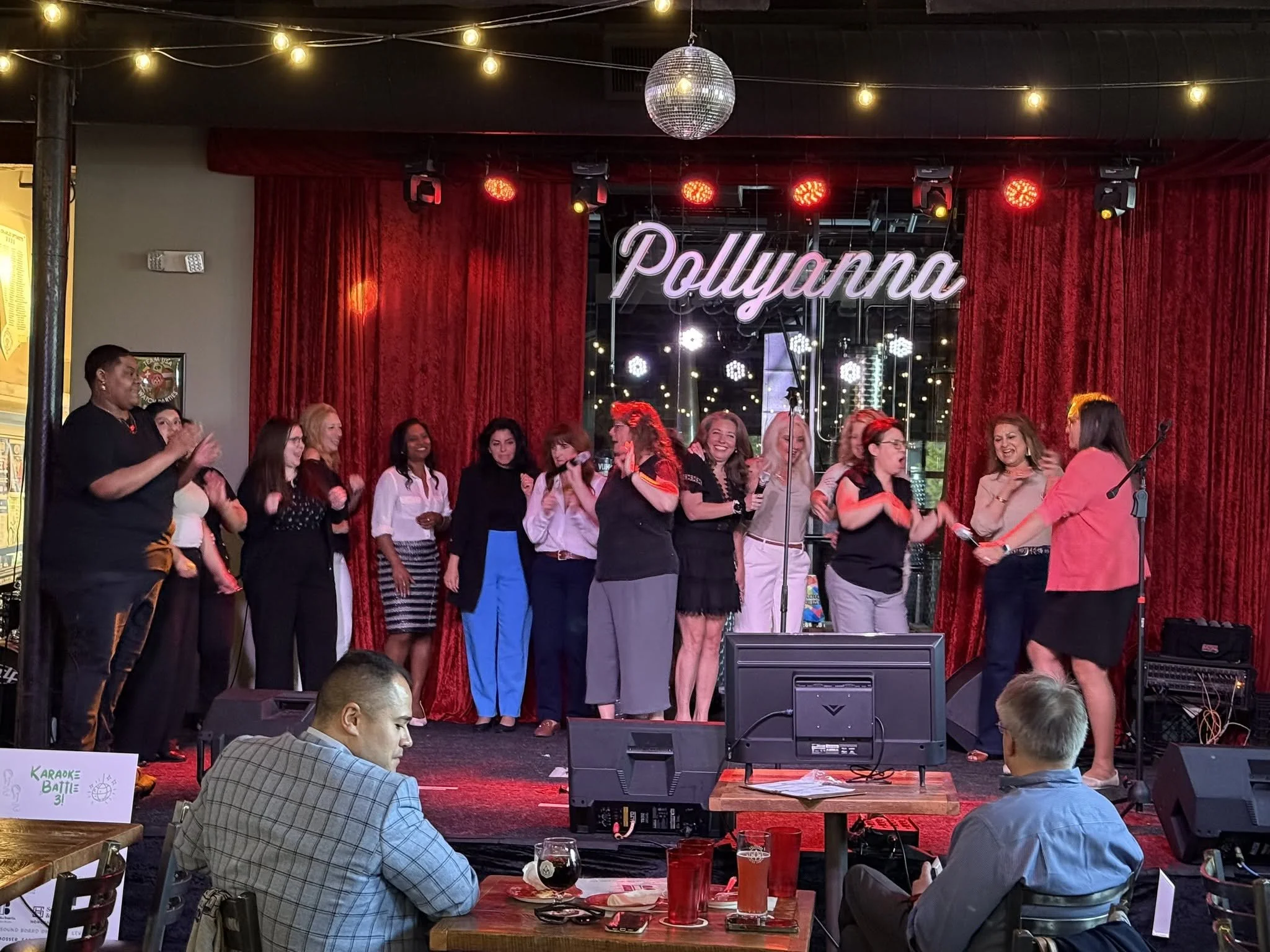 A group of women on stage singing or speaking into microphones, with red curtains, stage lighting, and a sign that reads 'Pollyanna.' There are people in the audience, some sitting at tables with drinks.