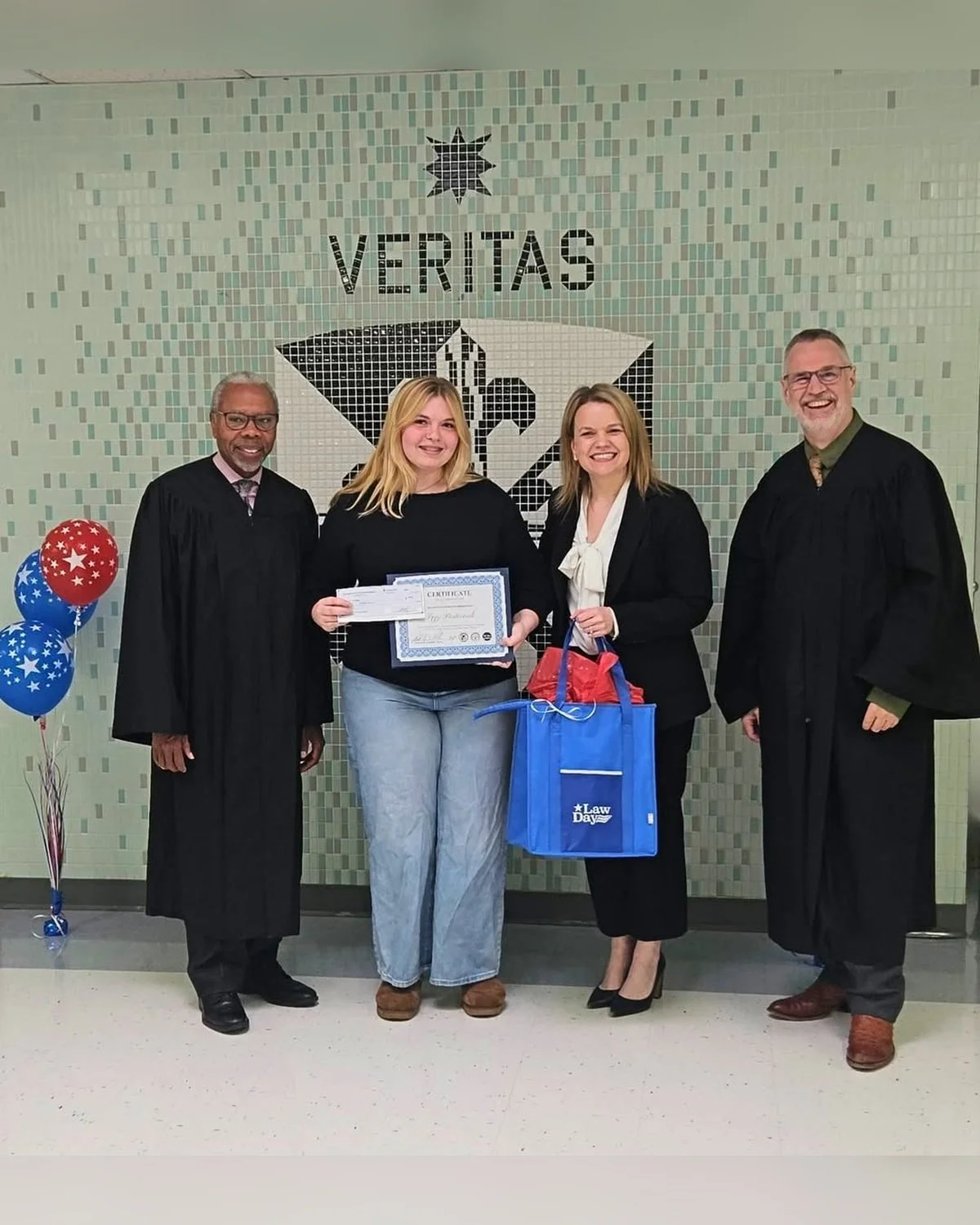 Group of four people standing in front of a mosaic wall with the word "VERITAS" and a symbol. The two women in the middle are holding a certificate and gifts, celebrating an achievement. Balloons on the left and a red and blue gift bag complete the scene.