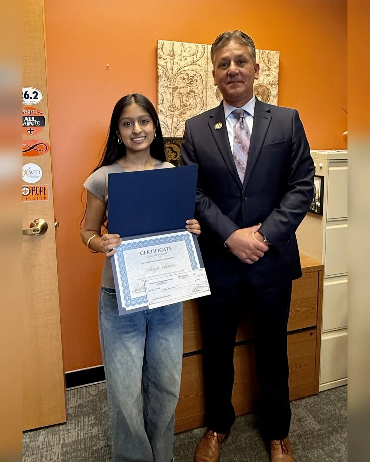 Young woman holding a certificate of achievement, standing next to a man in a suit, inside an office with orange walls and decorative artwork in the background.