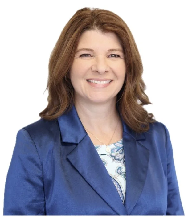 A woman with shoulder-length reddish-brown hair, smiling, wearing a blue blazer over a patterned blouse, against a white background.
