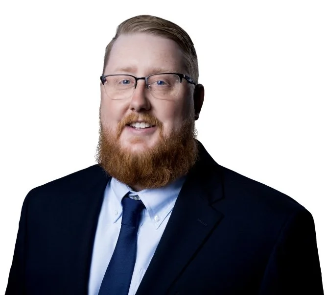 Portrait of a man with glasses, a red beard, wearing a suit and tie, smiling against a white background.