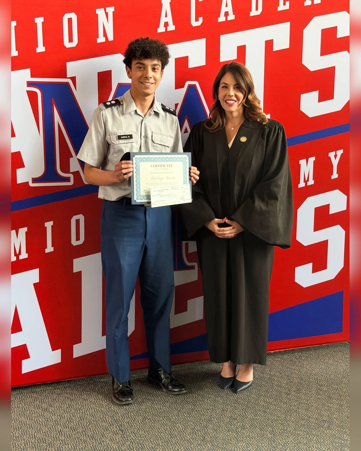 A young man in a uniform holding a certificate and a woman in a black robe standing on a red, white, and blue background, at an academic award event.