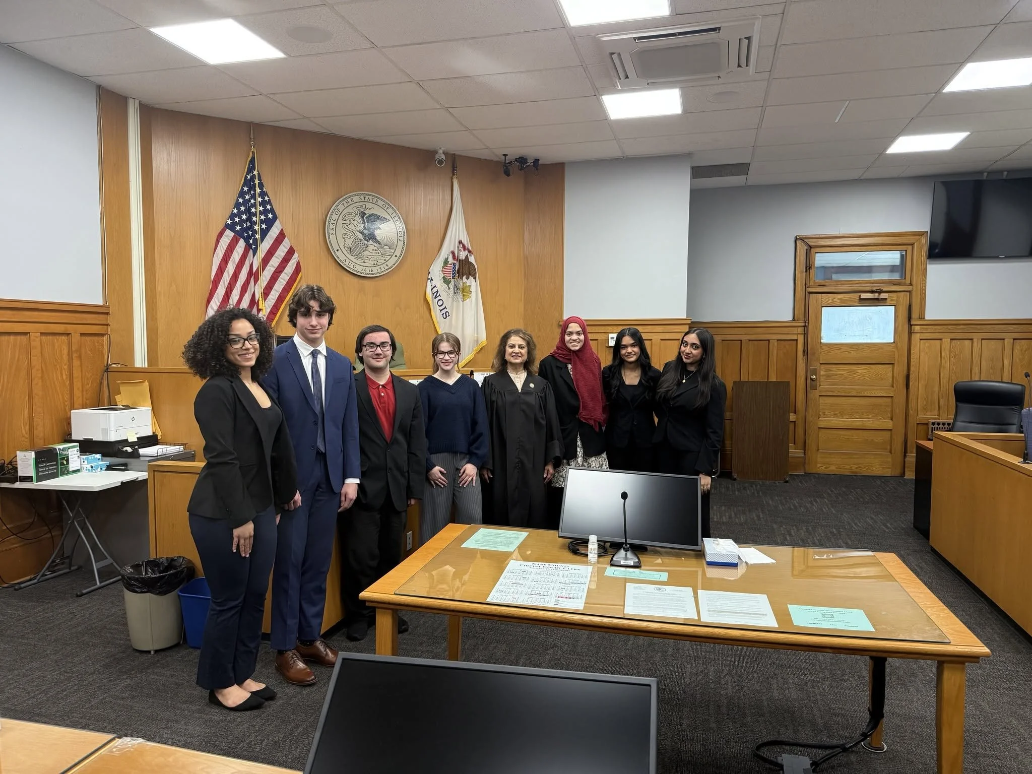 Group of six people in formal attire standing in a courtroom with wooden panels, American flag, Illinois state flag, and a seal on the wall behind them.