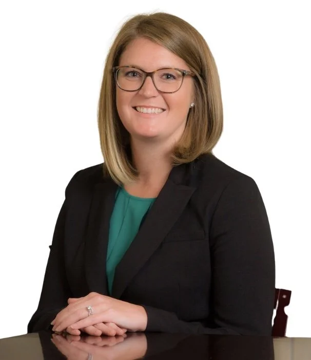 A woman with shoulder-length blonde hair, glasses, and a bright smile, wearing a black blazer and a teal blouse, sitting at a reflective surface with a white background.