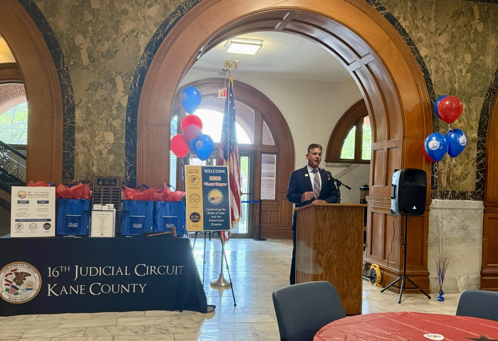A man in a dark suit and tie speaking at a podium during a formal event at the 16th Judicial Circuit in Kane County. The background features an American flag, a welcome sign, and a table with blue bags and informational materials. Red and blue balloons are arranged on the right side of the speaker, and the room has wooden arches and marble walls.