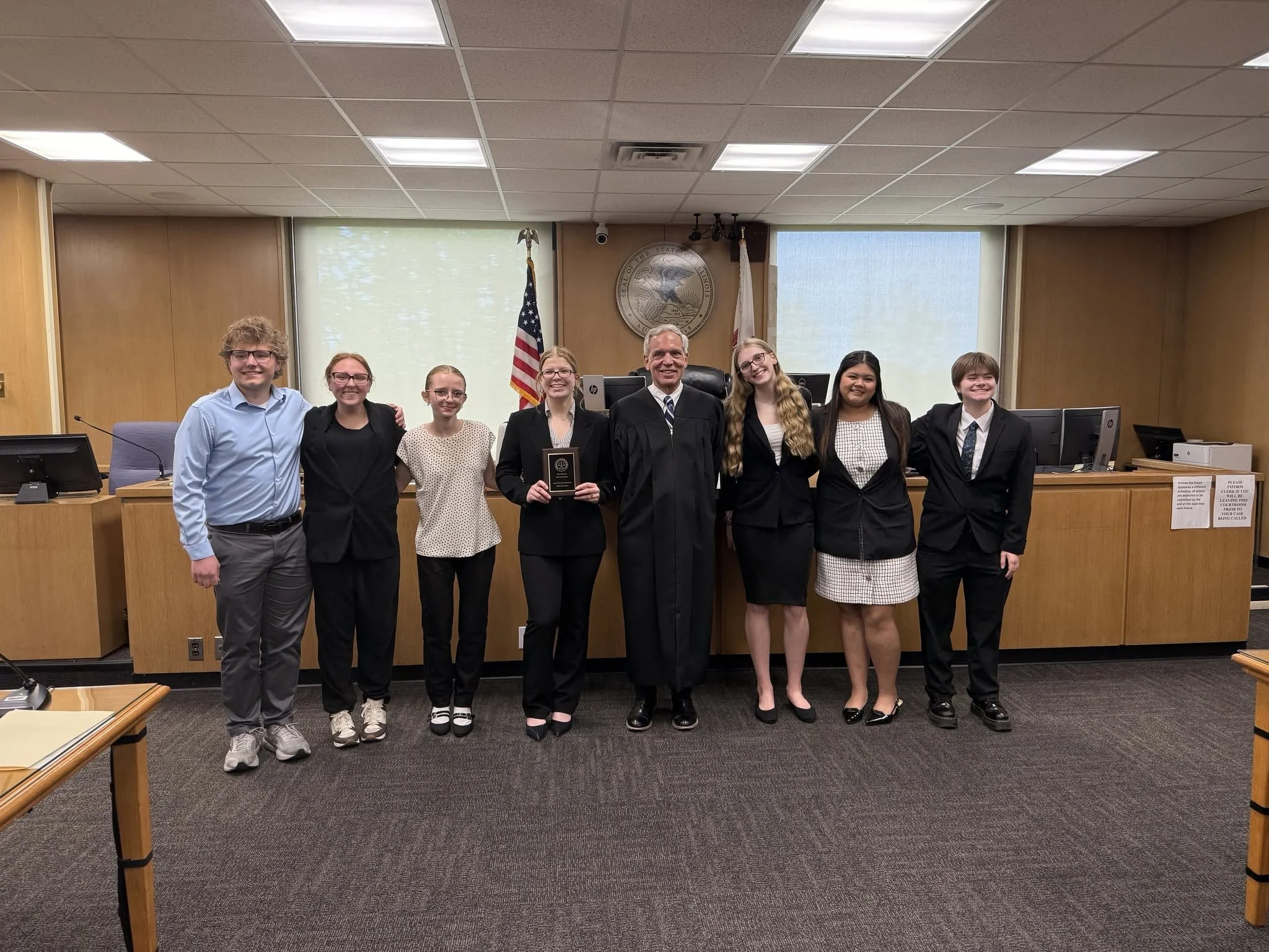 Group of eight people standing together in a courtroom, smiling at the camera, with a judge's bench, American flag, and state seal in the background.