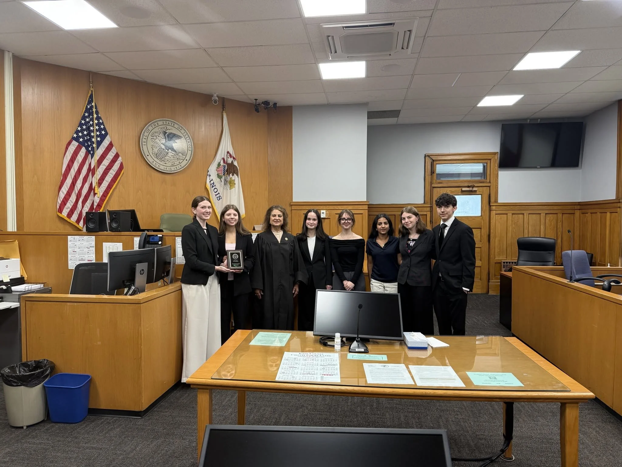 A group of seven young people and one woman in a black robe standing in a formal room with an American flag, a state flag, wood paneling, and a large flat screen TV on the wall. They are smiling, and one woman is holding a plaque.