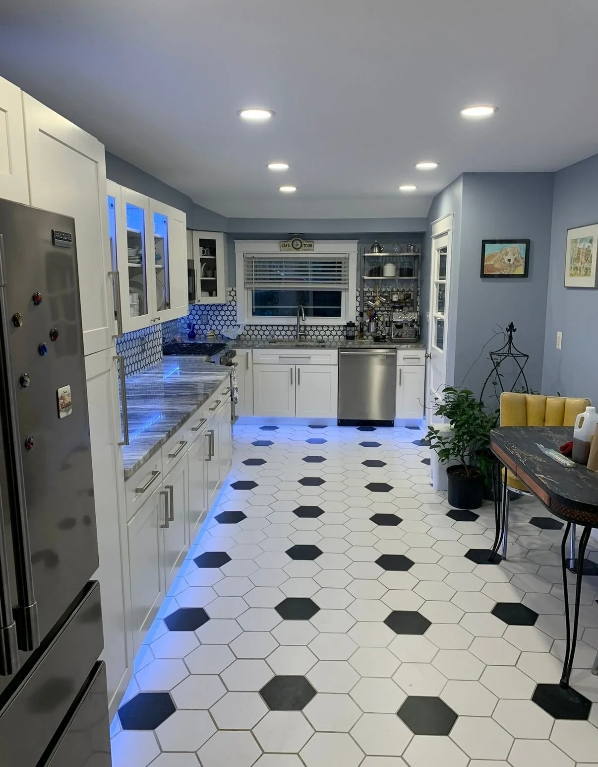 A modern kitchen with white cabinets, black and white hexagonal tile floors, stainless steel appliances, and blue LED lighting. There is a window with blinds, a small table with a potted plant, and artwork on the walls.