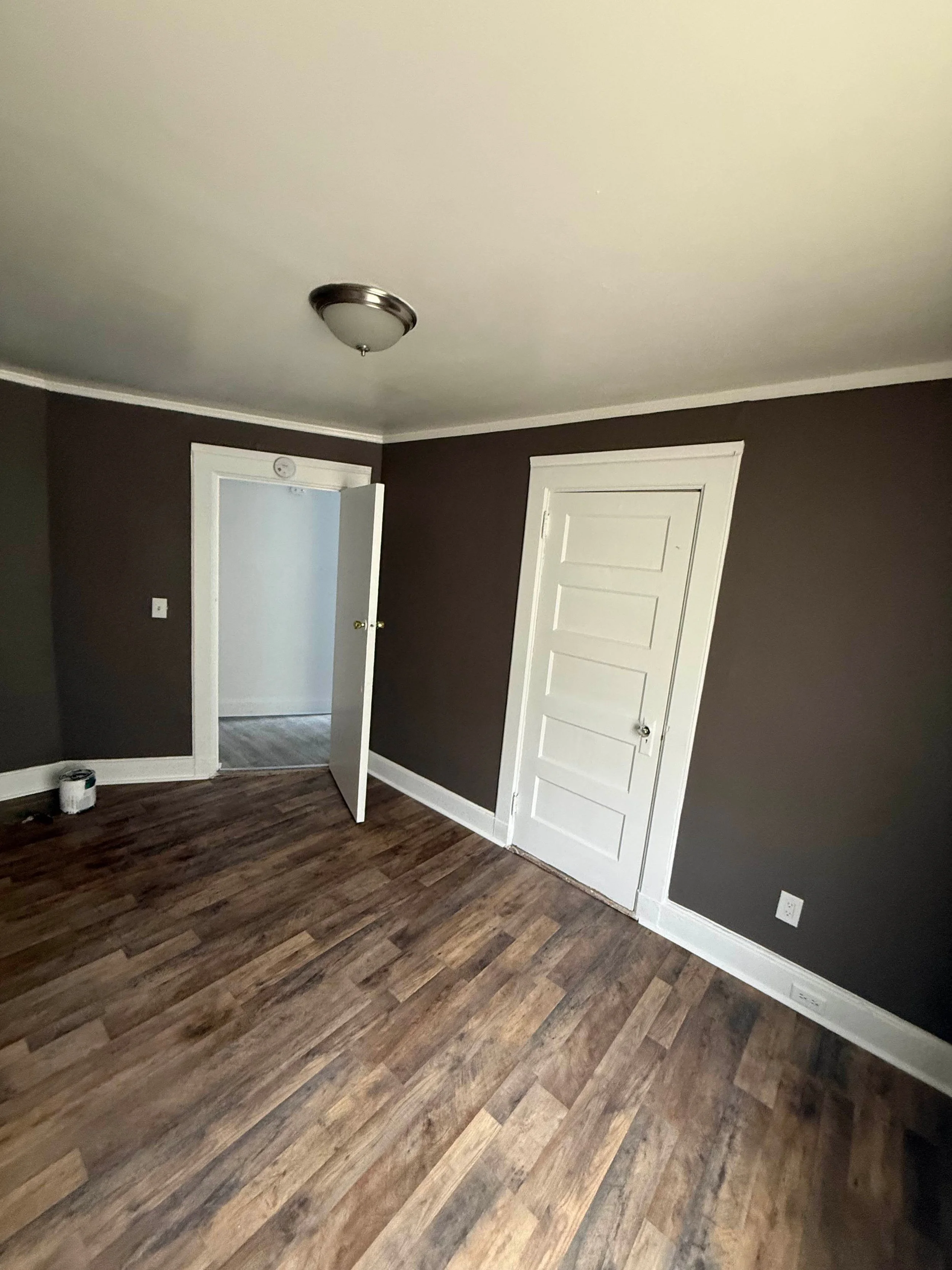 An empty room with dark brown wood flooring, dark brown walls, white trim, and two white doors, one of which is partially open.