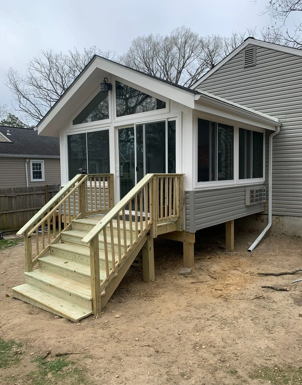 A newly constructed wooden staircase leading to a screened porch attached to a house. The house has grey vinyl siding and multiple large windows. The background shows neighboring houses and leafless trees, indicating a late fall or winter setting.