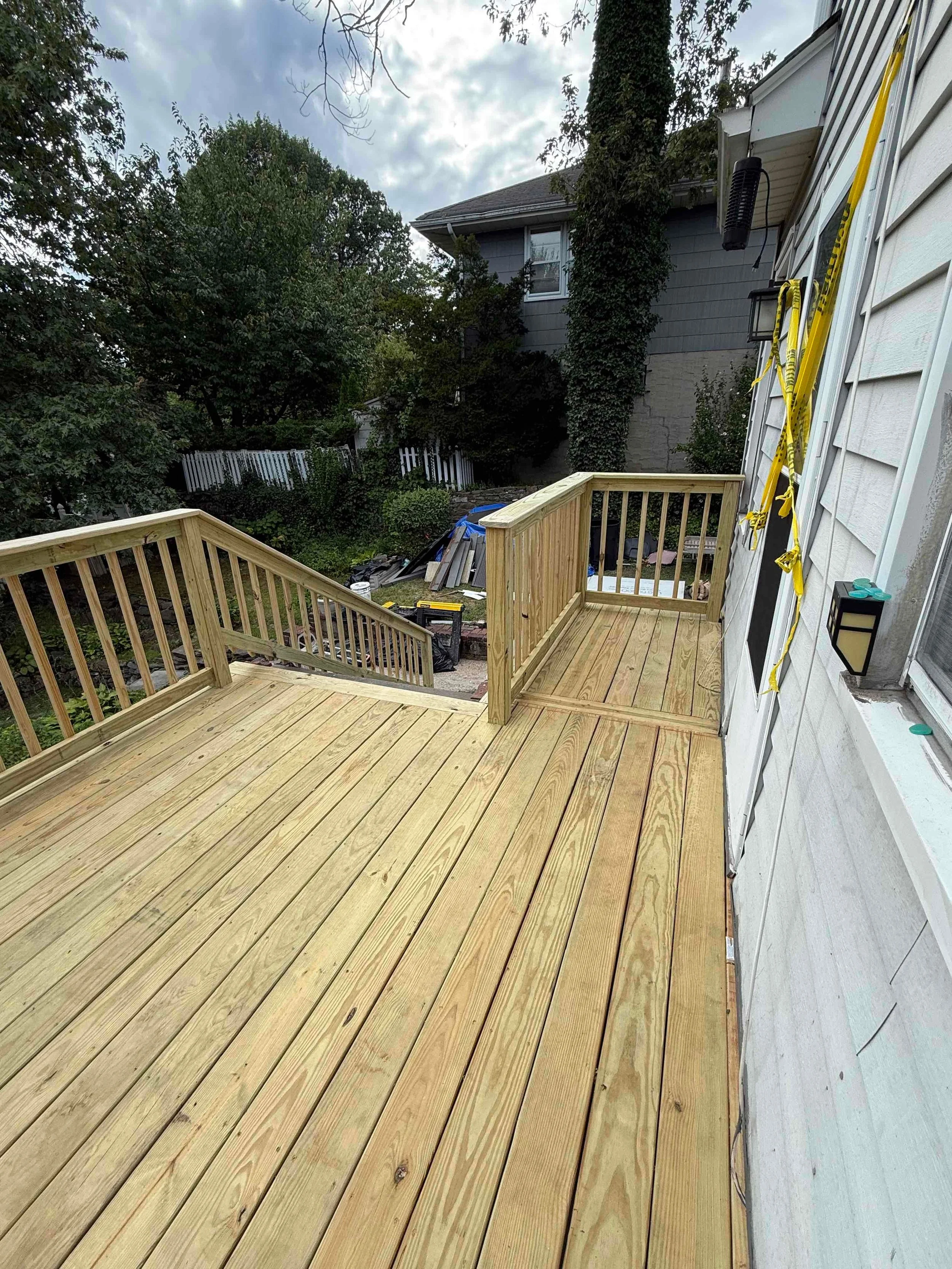 Newly built wooden deck with stairs, attached to a house with white siding and a black outdoor lantern, overlooking a backyard with trees, plants, and a white picket fence. Tools and construction materials are visible on the ground.