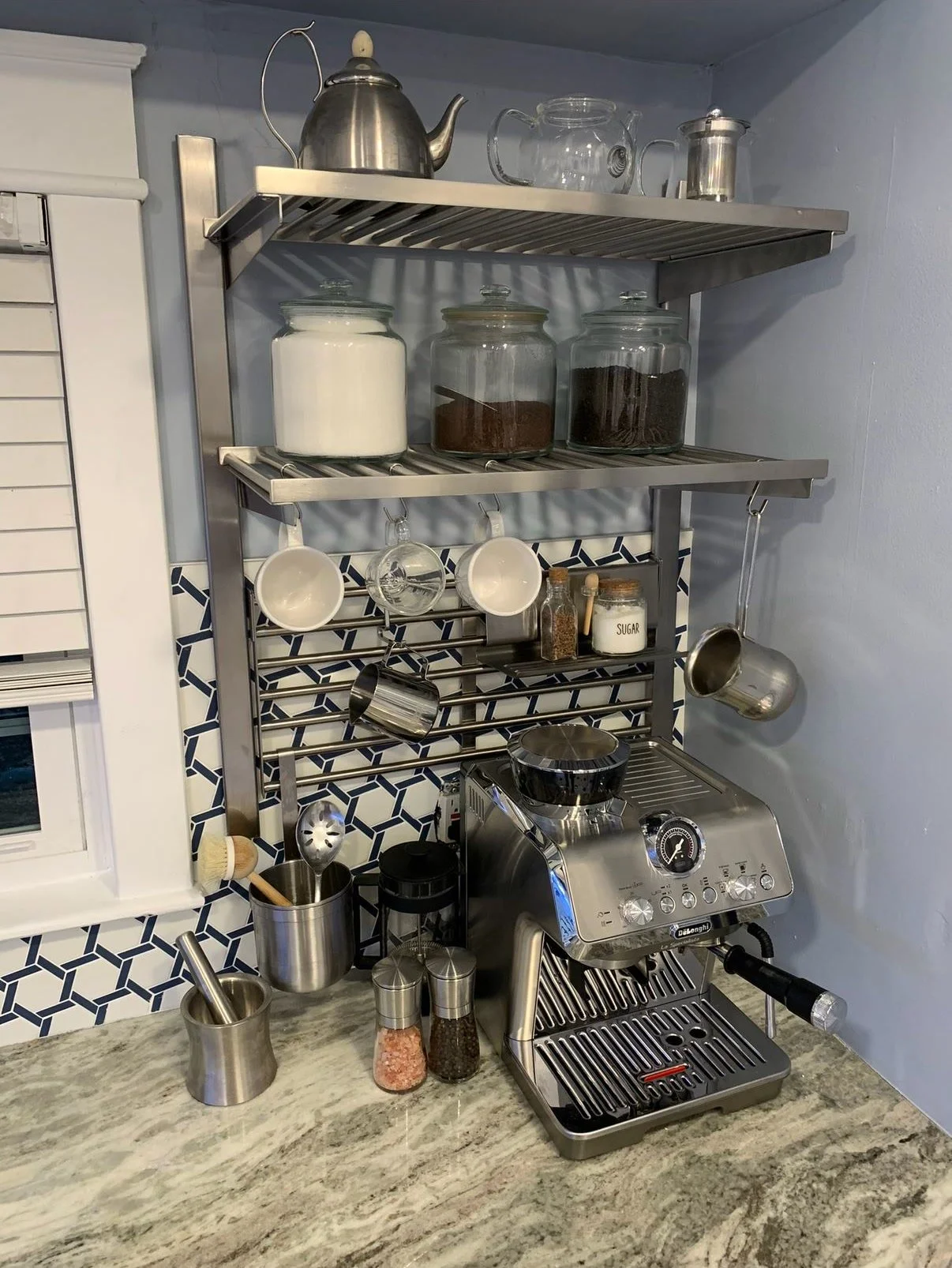 A kitchen countertop with a silver espresso machine, various utensils, jars of salt, sugar, and spices, and a stainless steel shelf holding teapots, cups, and jars of ingredients. There are mugs hanging on hooks and a patterned backsplash.