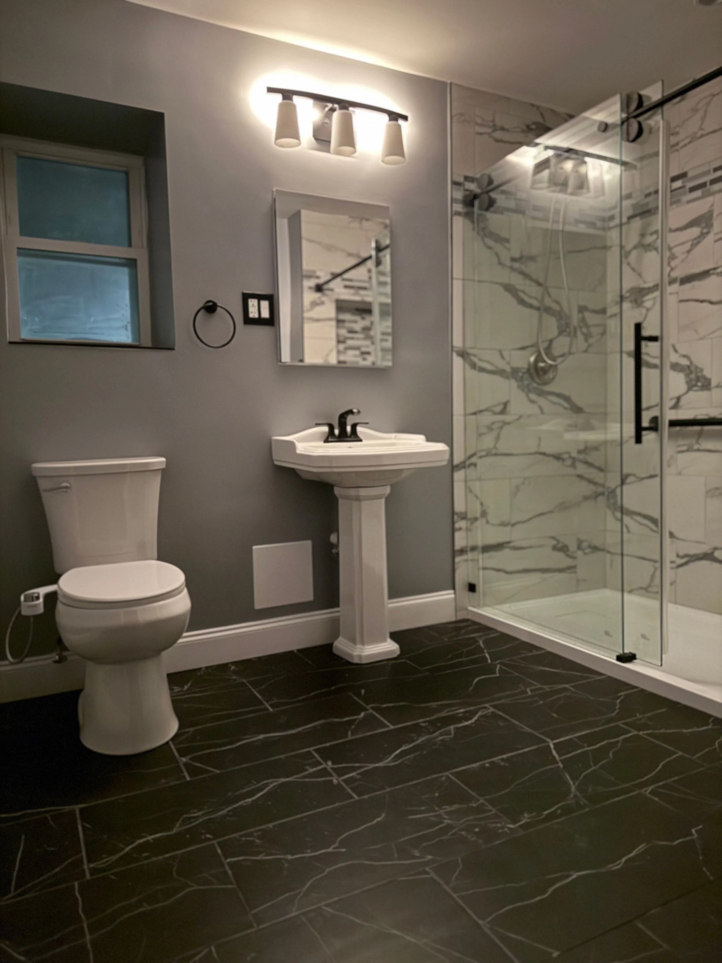 A modern bathroom featuring a toilet, a pedestal sink with a dark faucet, a mirror, a wall-mounted light fixture with three bulbs, a window, and a glass-enclosed shower with marble tile walls, black fixtures, and a black handle on the door. The floor