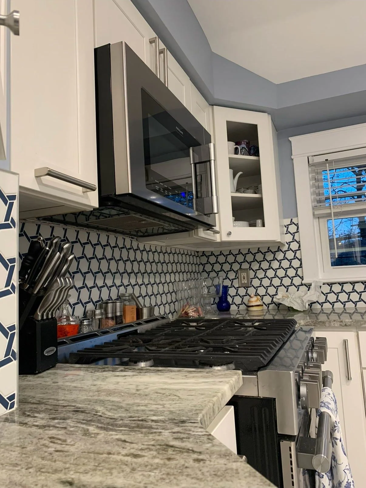 Kitchen stove with a granite countertop, knife holder, spice jars, microwave, open cabinet with dishes, window, and various kitchen items.
