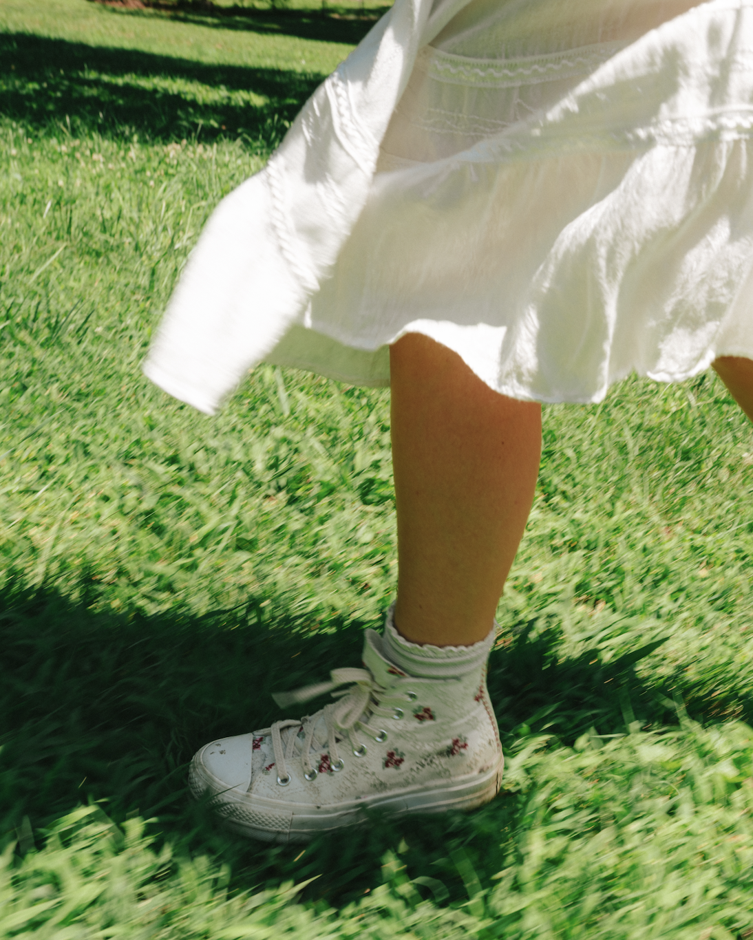 Close-up of a girl wearing white high-top sneakers with floral patterns, standing on grass with a white dress.