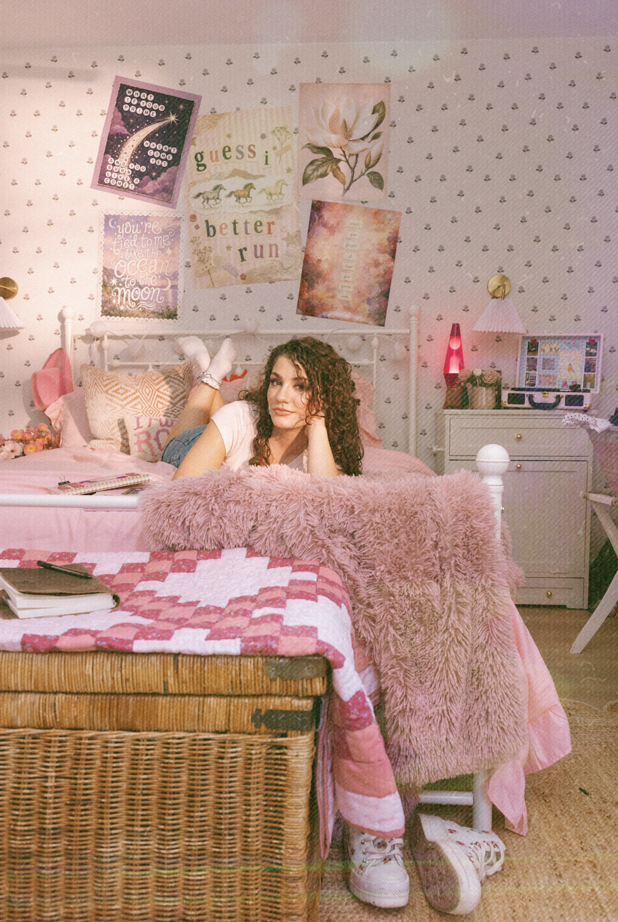A young woman with curly hair lying on her stomach on a pink bed with a fluffy pink blanket, in a pink-themed bedroom. The wall behind has various posters, including a moon, flowers, trees, and motivational quotes. There are bedside lamps, a book, a lava lamp, and a desk with a computer.