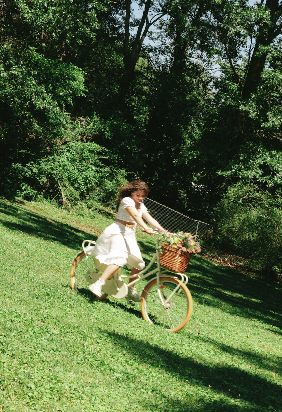 A girl riding a white bicycle with a wicker basket filled with flowers on a grassy field surrounded by green trees.