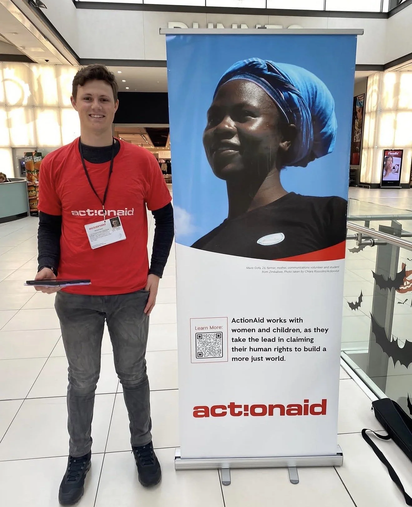 A young man with short brown hair wearing a red 'actionaid' T-shirt and a black lanyard stands next to a large, vertical promotional banner at an indoor location. The banner features a portrait of a woman wearing a blue headscarf, with text describing ActionAid's work with women and children to promote human rights. The background includes airport or mall-like decor, with a digital screen and decorative elements.