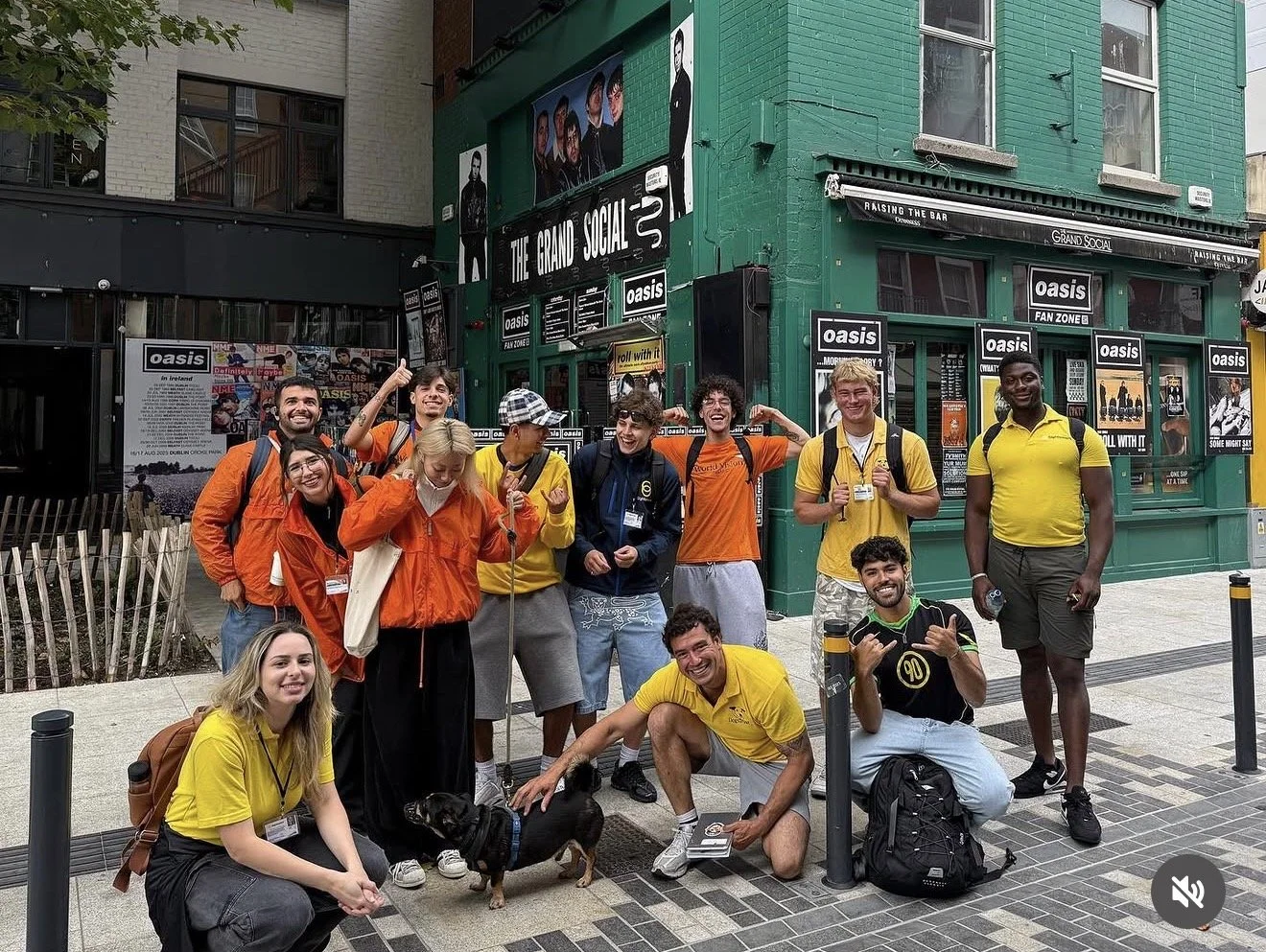 Group of people posing in front of a green building with posters and signs, including 'Oasis', 'The Grand Social', and 'Fan Zone', on a city sidewalk.