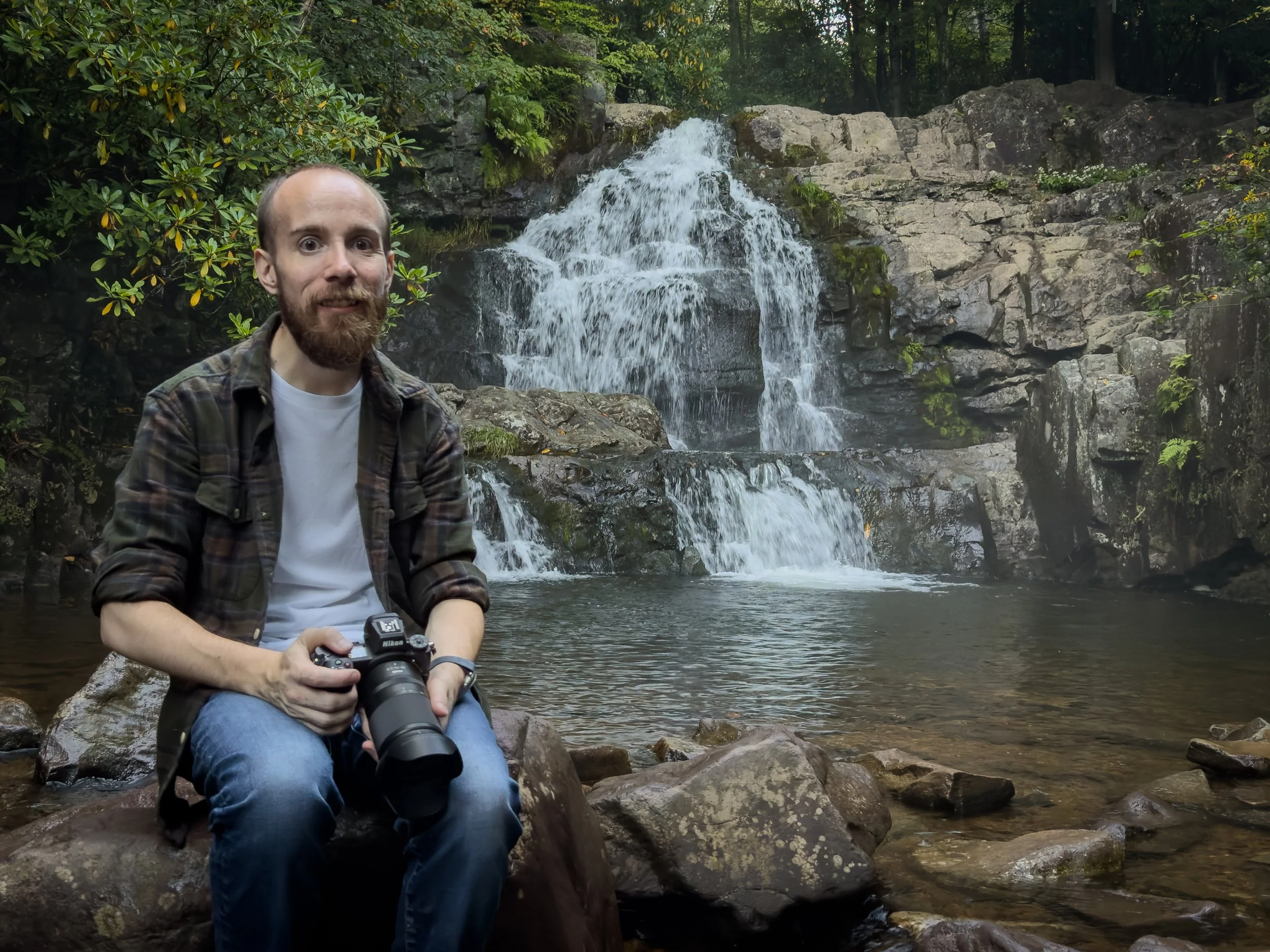 Shamus of O’Halligan Photography holding a camera beside a woodland waterfall.