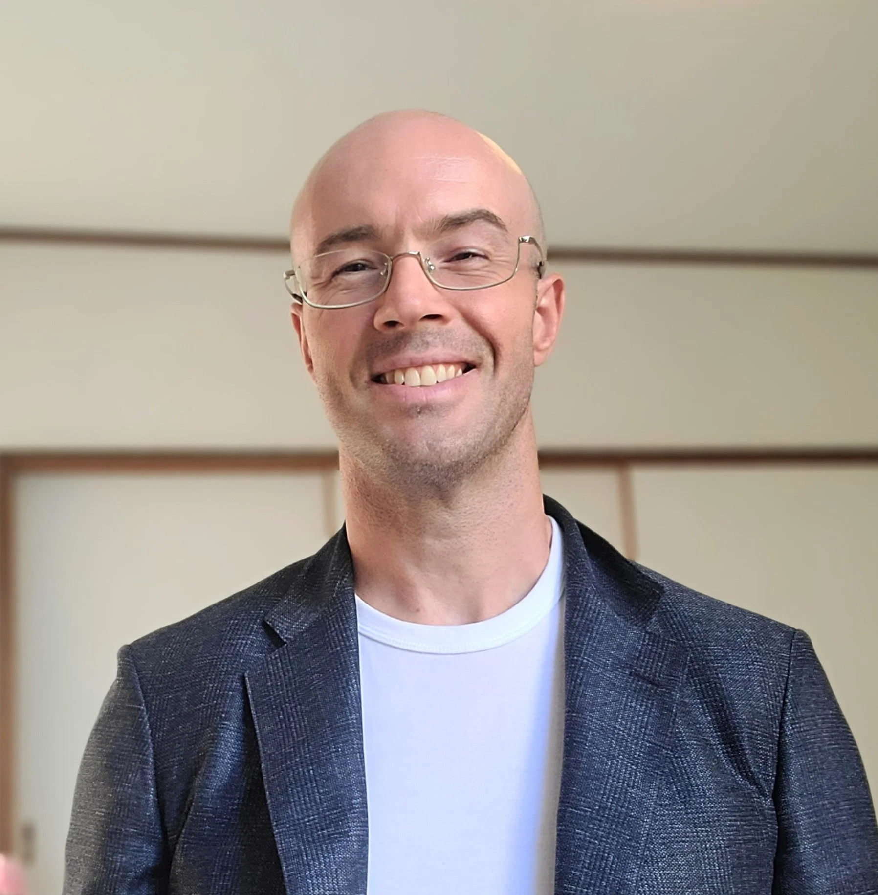 Smiling man with glasses and bald head, wearing a gray blazer over a white shirt, standing against a beige wall.