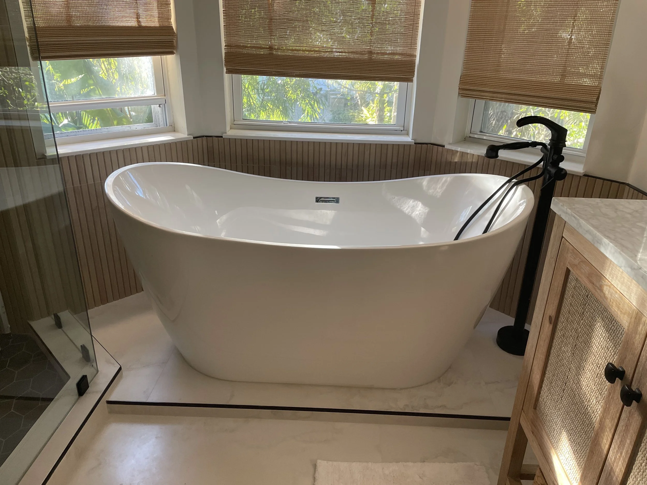 A bathroom with a large bathtub positioned in front of three windows with wooden blinds. There is a faucet and handheld shower attachment mounted on the right side of the tub, and the bathroom has a beige tile floor and wooden paneling on the lower walls.