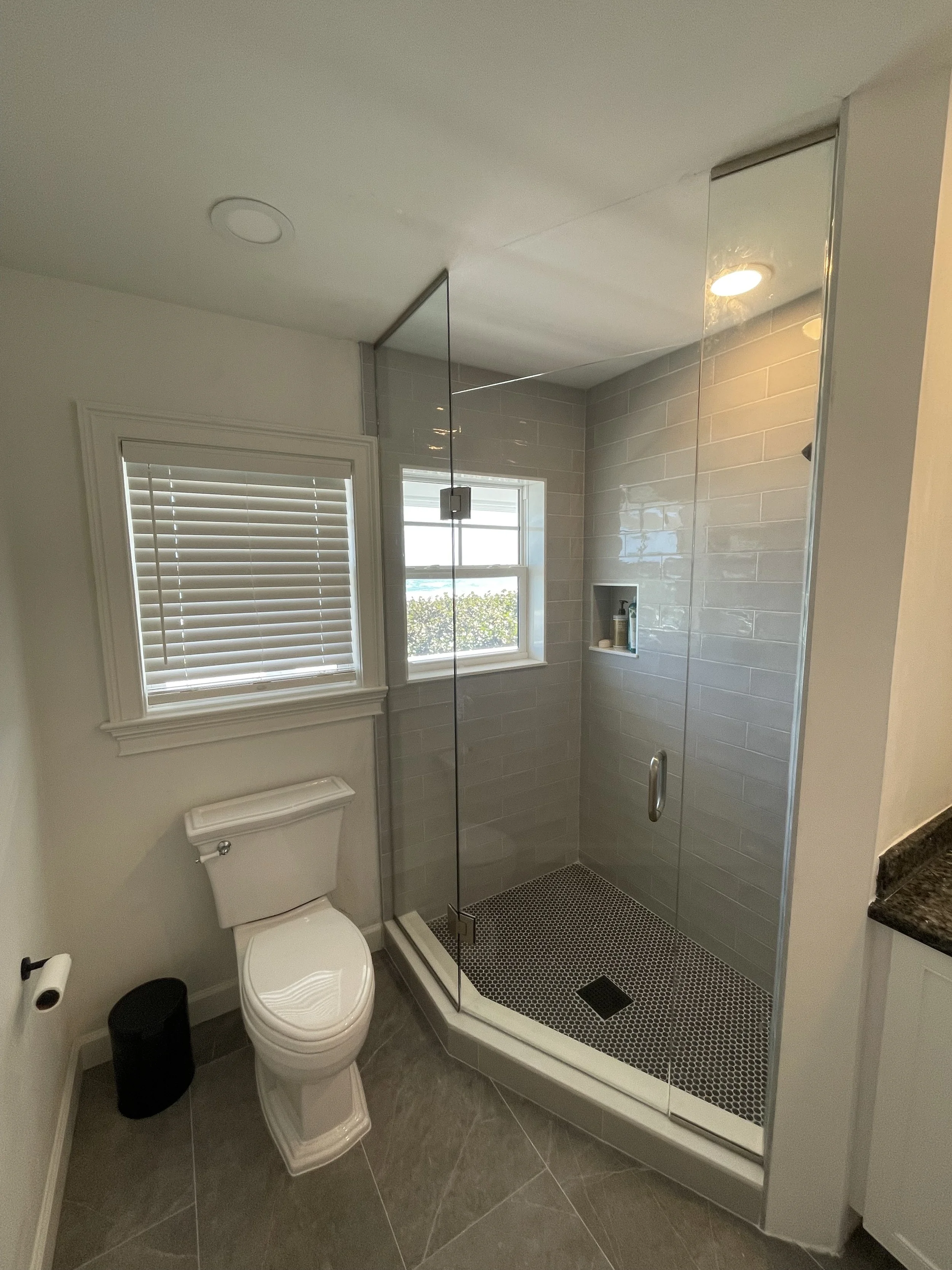 A bathroom featuring a toilet, a window with blinds, and a glass-enclosed walk-in shower with gray tile walls and black pebble floor tiles.