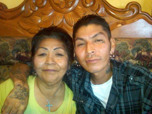 A young man with tattoos, earrings, and a checkered shirt taking a selfie with an older woman wearing a yellow top and a cross necklace, sitting on a wooden headboard with a decorative panel and a patterned wall behind them.