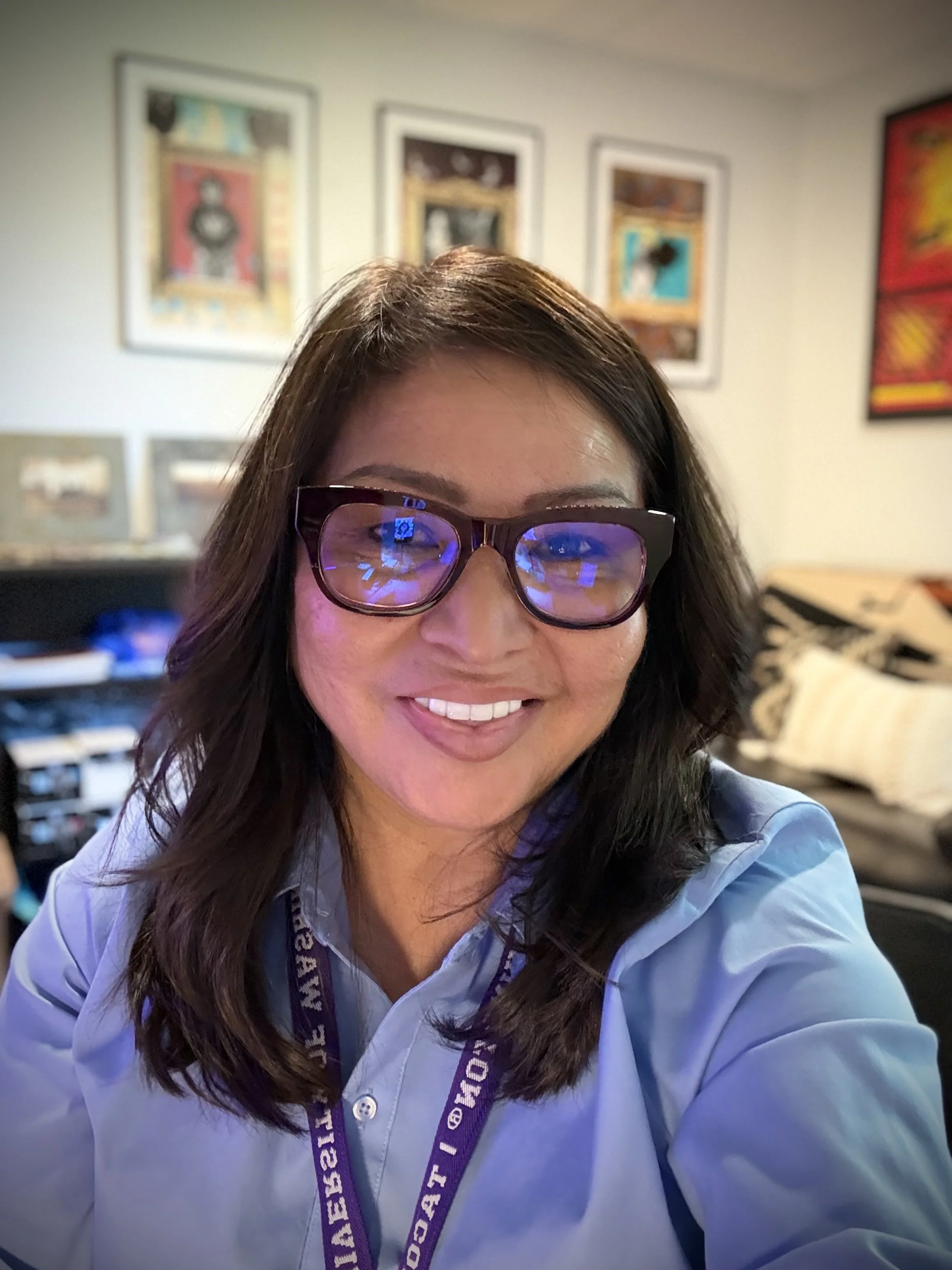 A woman with long brown hair wearing glasses and a light blue collared shirt, smiling indoors with framed artwork on the wall behind her.