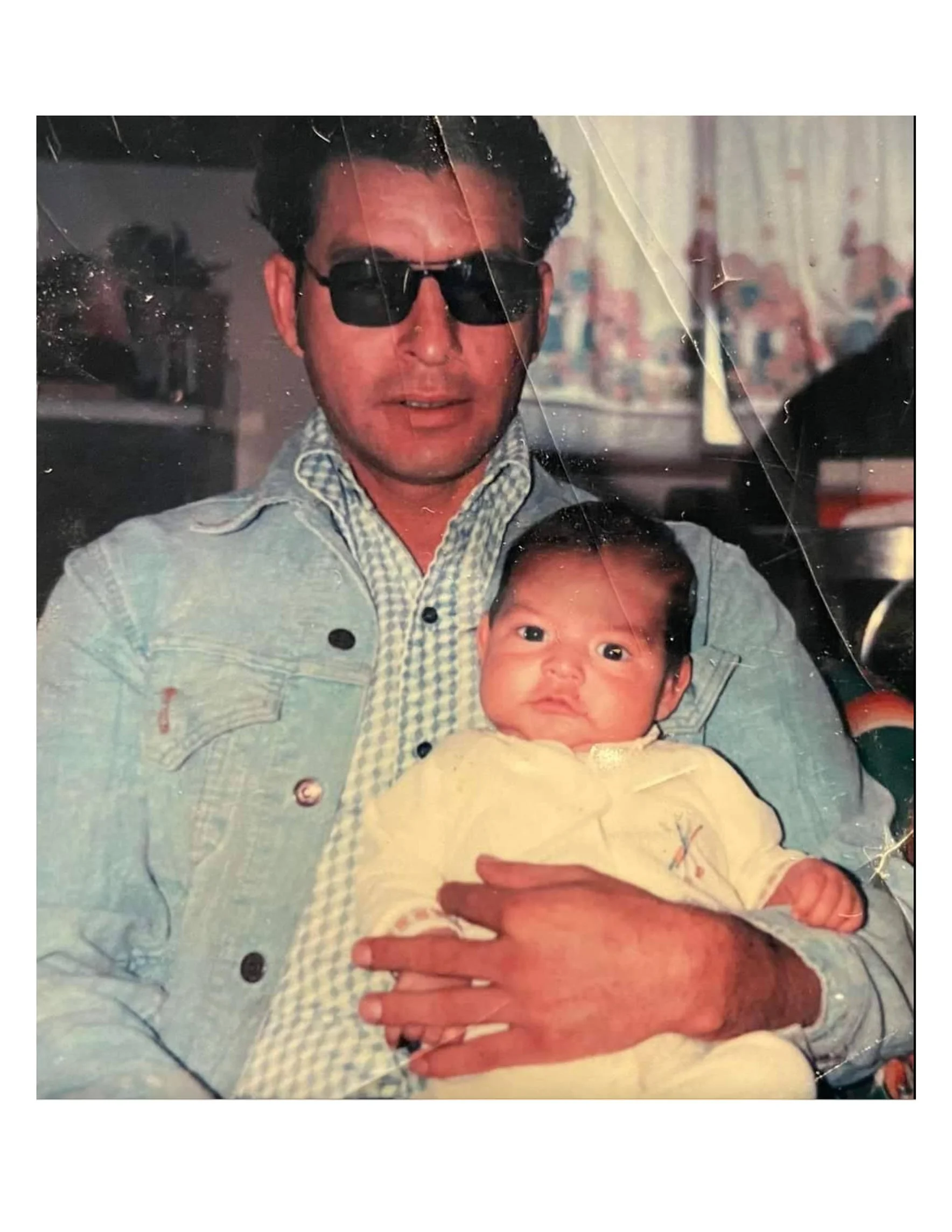 A man wearing sunglasses and a denim jacket holding a baby with wide eyes and a yellow outfit, in an indoor setting with a blurred background.