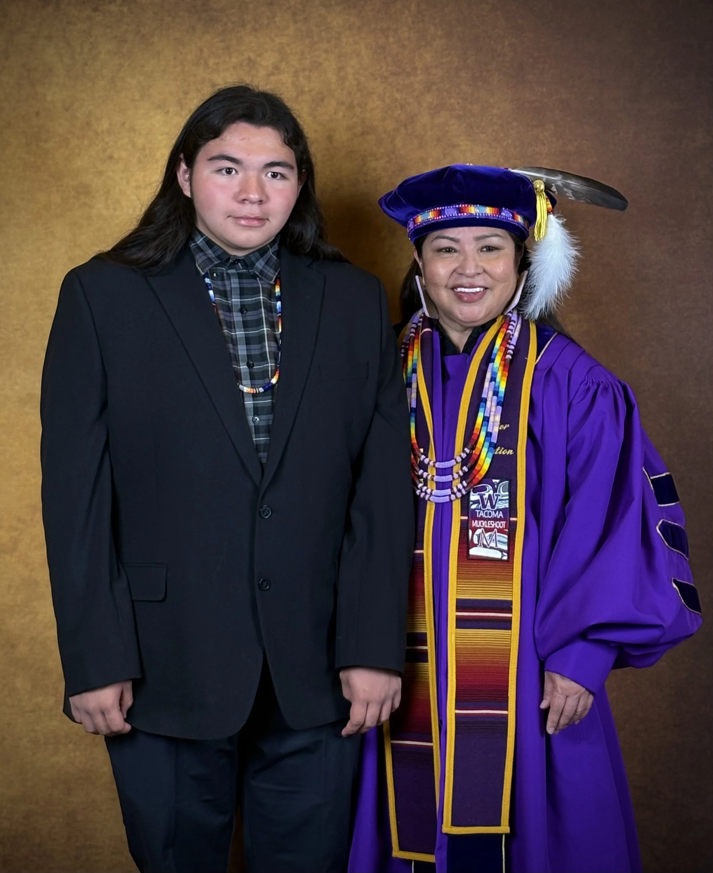 Young man in a dark suit and plaid shirt standing next to woman in graduation regalia, including a purple gown and a decorated doctoral cap with feathers, against a gold background.