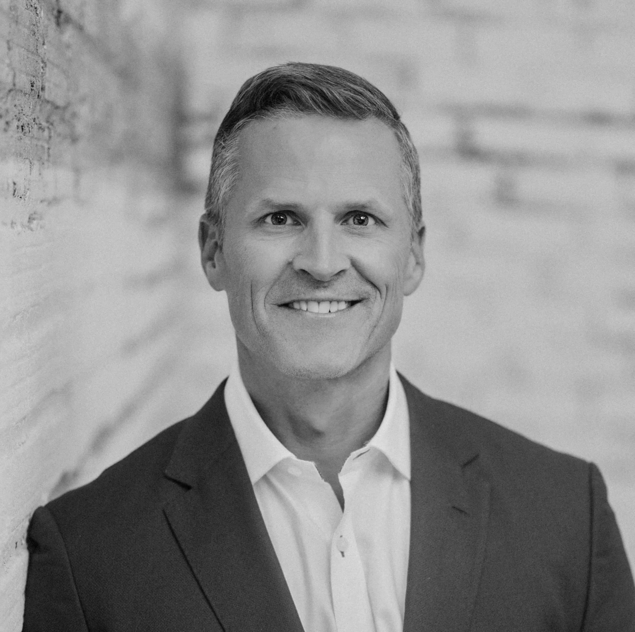 Black and white photo of a middle-aged man with short hair, dressed in a suit, smiling, standing outdoors near a brick wall.
