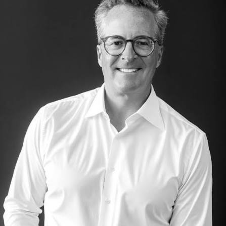 Portrait of a smiling man with glasses, wearing a white shirt, against a dark background.