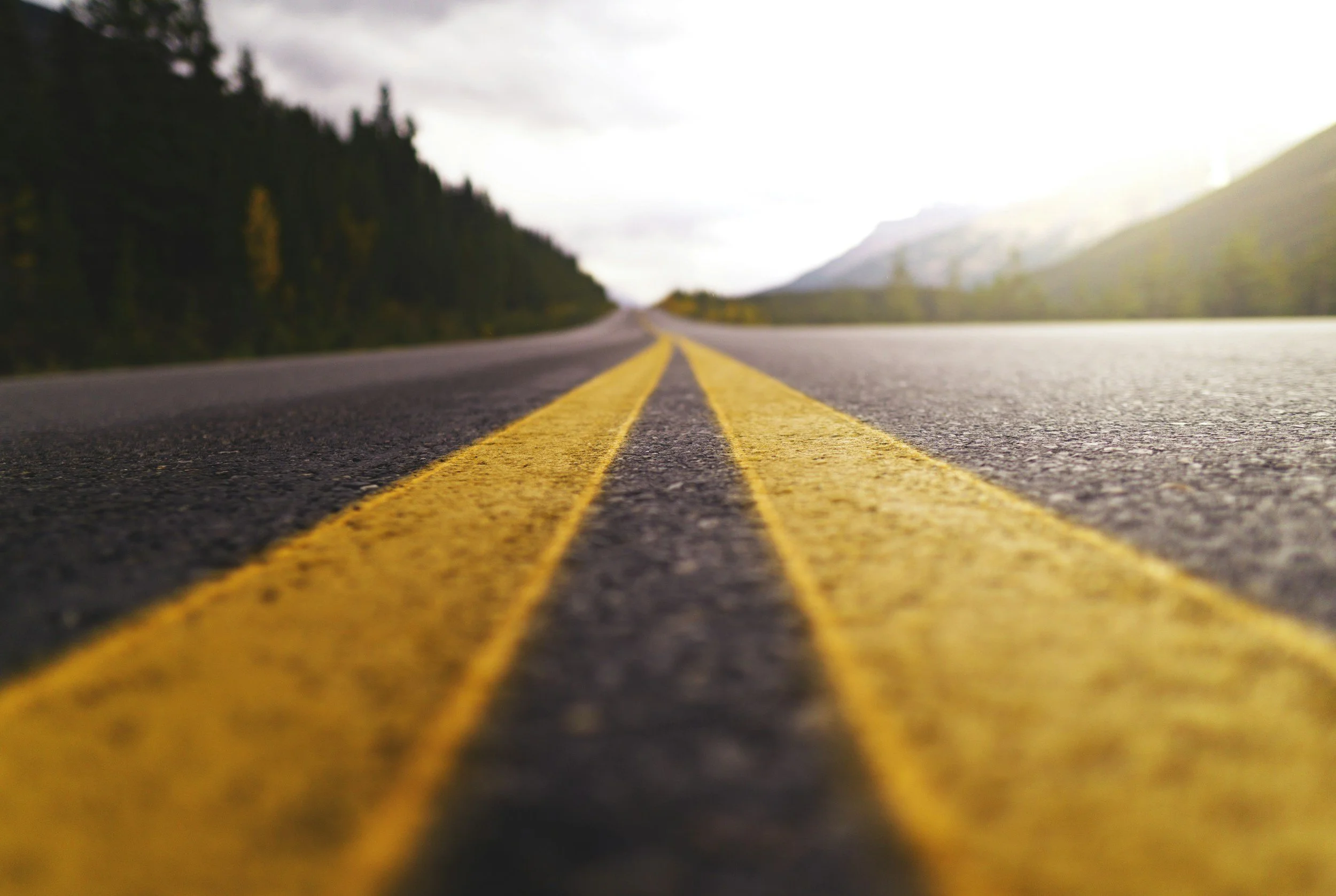 A close-up view of an empty two-lane road with yellow dividing lines, surrounded by trees and mountains under an overcast sky.